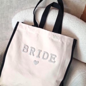May include: A canvas tote bag with black trim and handles. The word "BRIDE" is spelled out in sparkly silver letters, with a small silver heart below. The bag is off-white and sits on a light-colored surface.
