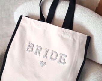May include: A canvas tote bag with black trim and handles. The word "BRIDE" is spelled out in sparkly silver letters, with a small silver heart below. The bag is off-white and sits on a light-colored surface.