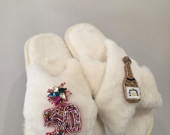 May include: A pair of white faux fur slippers featuring decorative patches. The left slipper has a pink beaded number 50 with a colorful crystal starburst above it. The right slipper features a beaded champagne bottle patch with the text Champagne.
