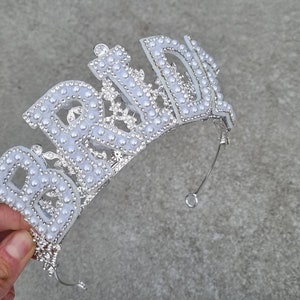 May include: A silver tiara with the word "Bride" spelled out in white plastic letters and decorated with white pearl-like beads.