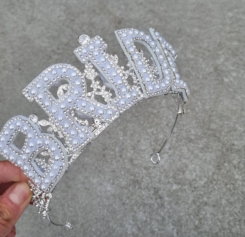 May include: A silver tiara with the word "Bride" spelled out in white plastic letters and decorated with white pearl-like beads.