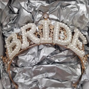 May include: A gold-toned tiara with the word "BRIDE" spelled out in white letters with pearl accents. The tiara is decorated with small rhinestones and pearls.
