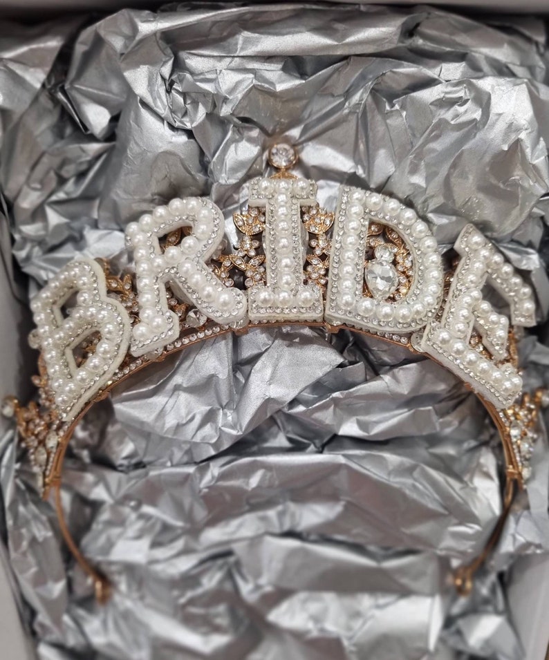 May include: A gold-toned tiara with the word "BRIDE" spelled out in white letters with pearl accents. The tiara is decorated with small rhinestones and pearls.