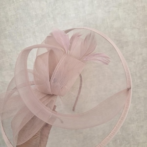 May include: A pale pink fascinator with a circular base and a delicate mesh overlay. The headpiece features a large bow and several feathers, all in matching shades of pink. The design is elegant and suitable for formal events.