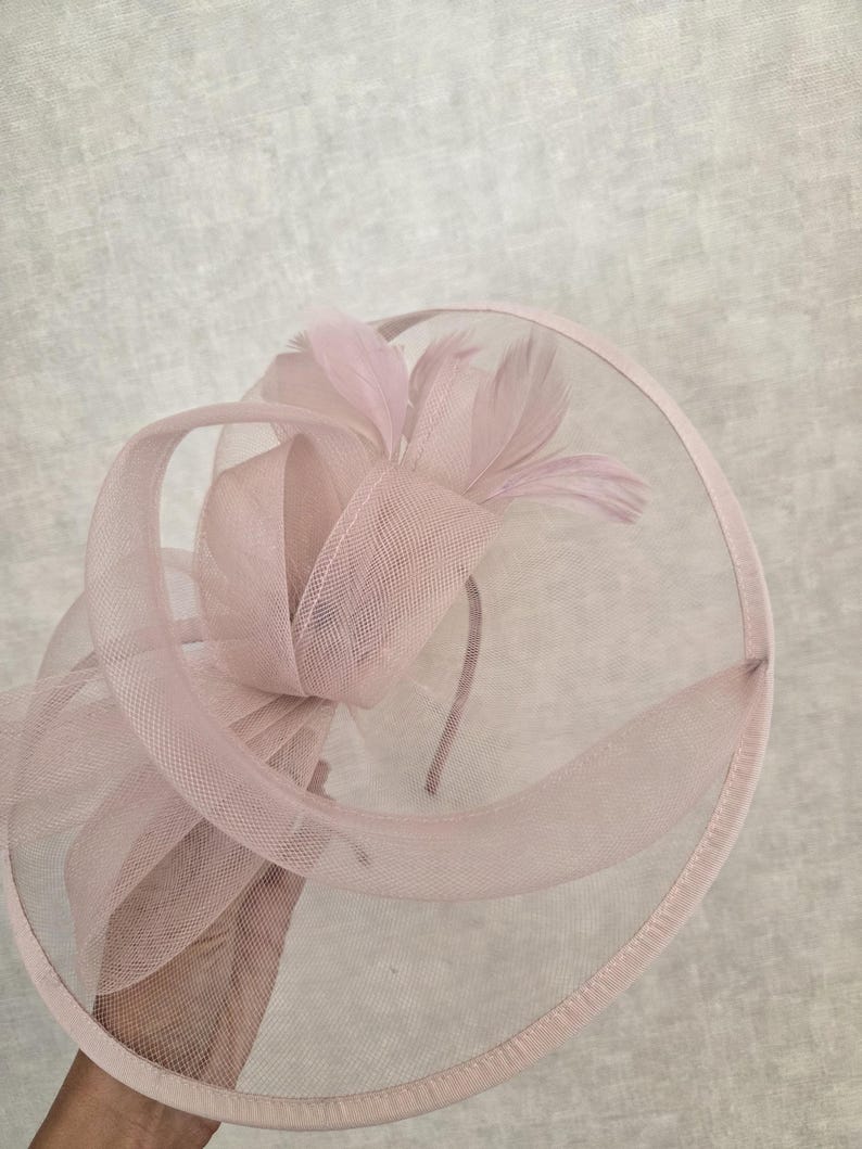 May include: A pale pink fascinator with a circular base and a delicate mesh overlay. The headpiece features a large bow and several feathers, all in matching shades of pink. The design is elegant and suitable for formal events.