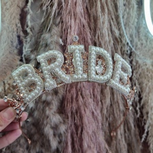 May include: A gold-toned tiara with the word "BRIDE" spelled out in white pearls and rhinestones. The tiara has a delicate, feminine design.
