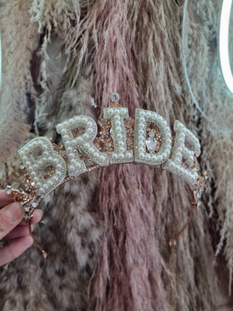 May include: A gold-toned tiara with the word "BRIDE" spelled out in white pearls and rhinestones. The tiara has a delicate, feminine design.