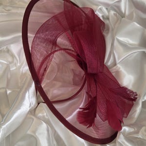 May include: A burgundy-colored formal hat with a wide brim and decorative bow. The hat features a mesh design and is adorned with feathers. The hat is likely for a special occasion.