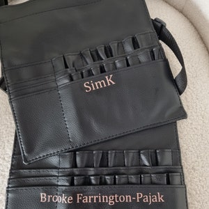 May include: Black leather makeup brush roll with multiple pockets and a strap. The roll has two personalized names, "SimK" and "Brooke Farrington-Pajak".