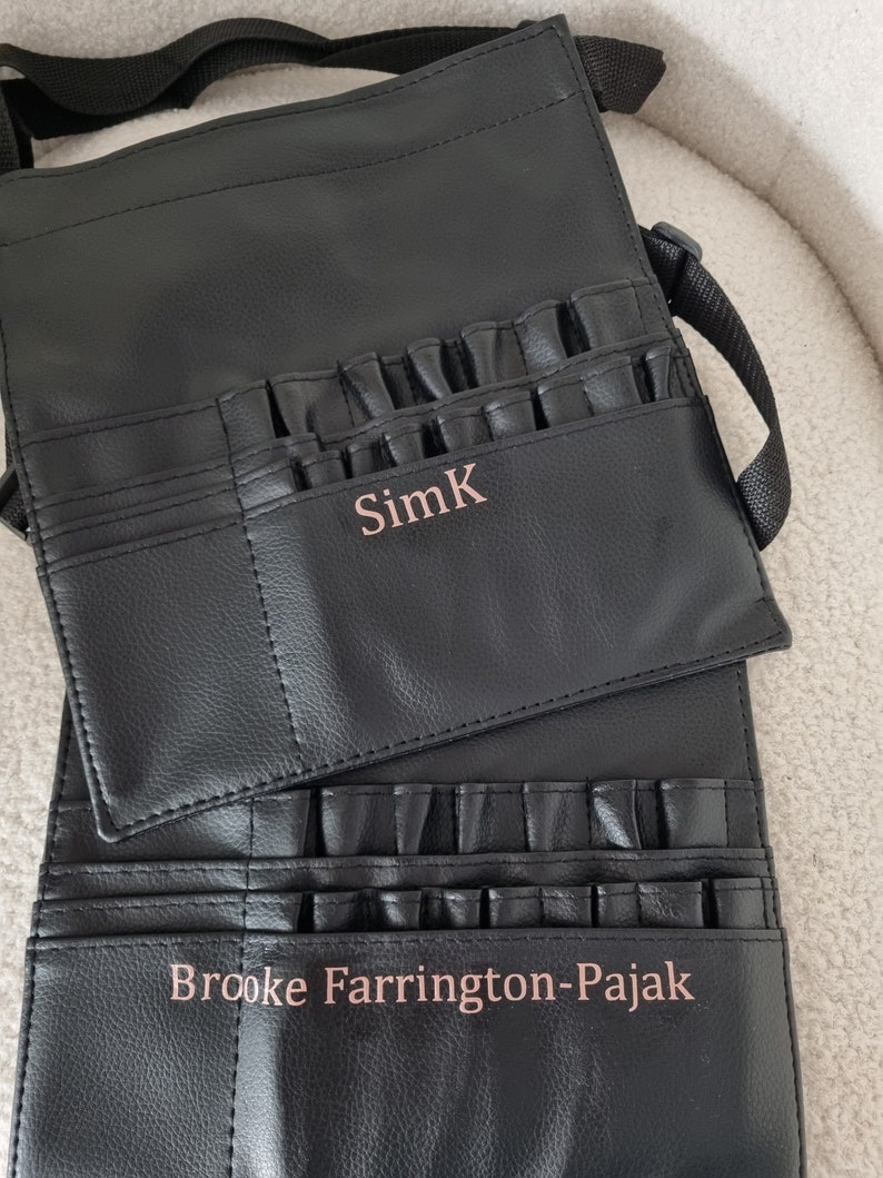 May include: Black leather makeup brush roll with multiple pockets and a strap. The roll has two personalized names, "SimK" and "Brooke Farrington-Pajak".