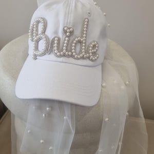 K&ouml;nnte beinhalten: Wei&szlig;e Baseballkappe mit einem mit Perlen besetzten "Bride"-Design und einem daran befestigten wei&szlig;en T&uuml;llschleier.