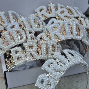 May include: A set of white and gold bridal headbands with the word "Bride" spelled out in pearl-like beads. The headbands are in a white box.