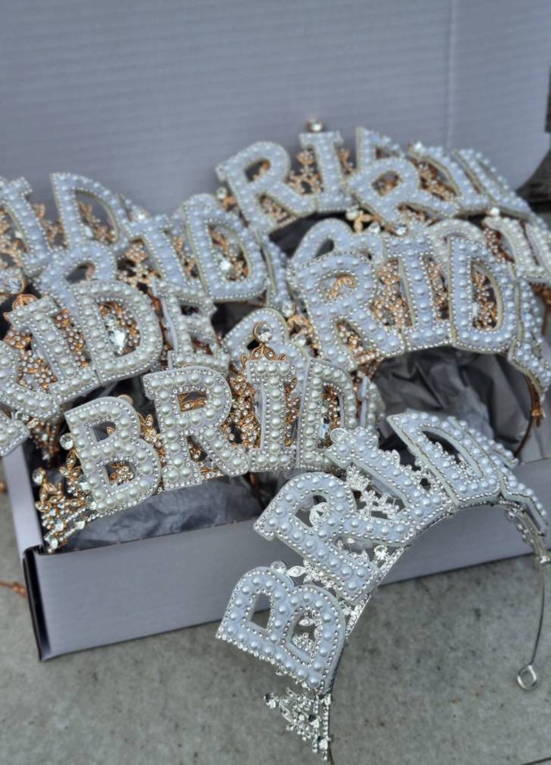 May include: A set of white and gold bridal headbands with the word "Bride" spelled out in pearl-like beads. The headbands are in a white box.