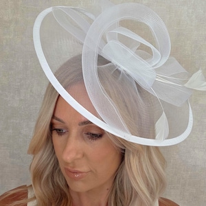 May include: A white fascinator hat with a circular brim and a decorative swirl design. The hat features a mesh overlay and is adorned with feathers. This elegant headpiece is suitable for special occasions.