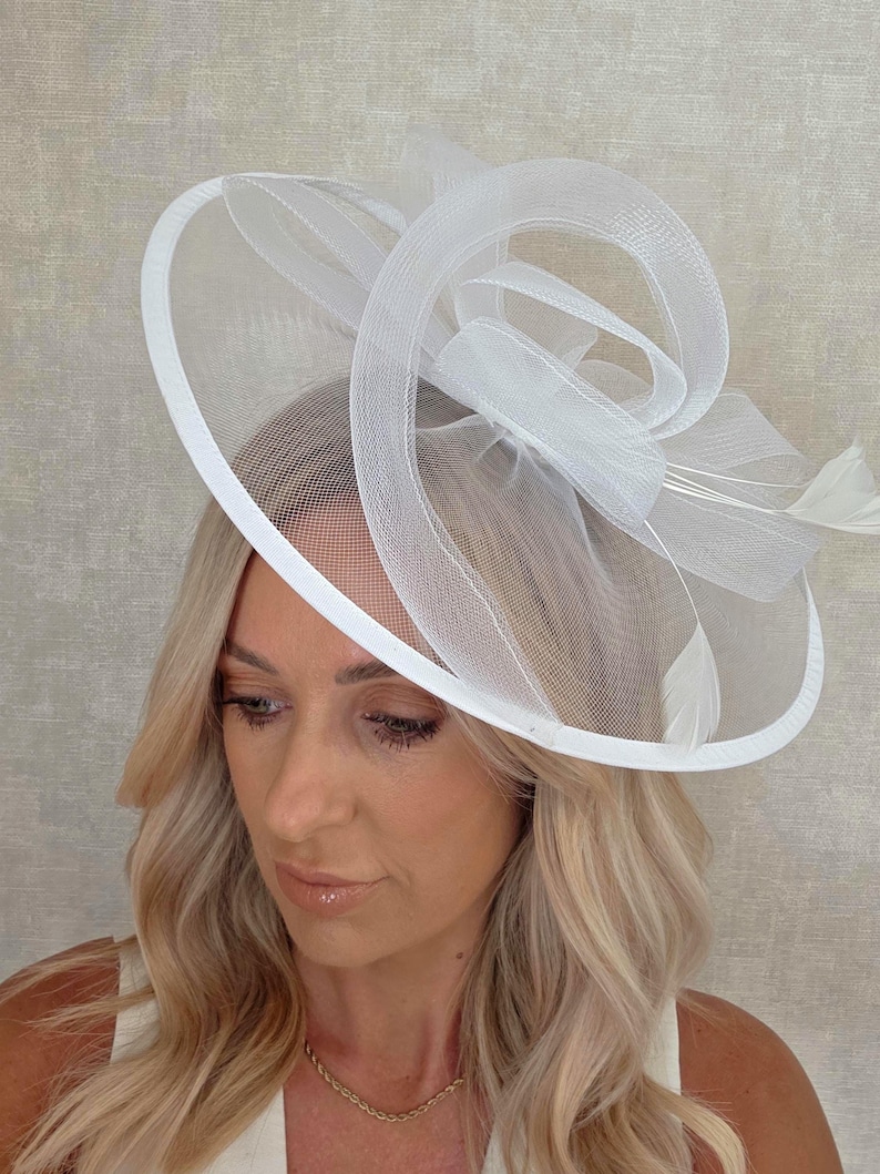 May include: A white fascinator hat with a circular brim and a decorative swirl design. The hat features a mesh overlay and is adorned with feathers. This elegant headpiece is suitable for special occasions.