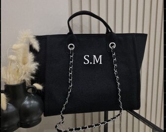 May include: Black tote bag with silver chain straps and handles. The bag features the initials "S.M" in white. The bag is sitting on a glass surface.