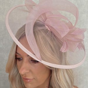 May include: A light pink formal hat with a wide brim and decorative mesh overlay. The hat features a large bow and feather accents, suitable for special occasions. The hat is a soft, pastel pink color.