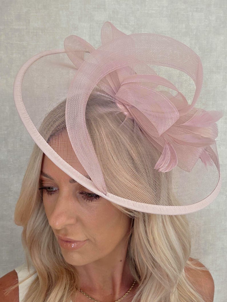 May include: A light pink formal hat with a wide brim and decorative mesh overlay. The hat features a large bow and feather accents, suitable for special occasions. The hat is a soft, pastel pink color.