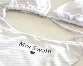 May include: White ribbed bikini set. The top is a triangle style with adjustable ties. The bottom features the text "Mrs Swain" with a black heart. The set is made of a textured fabric.