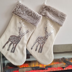 May include: Two cream-colored Christmas stockings with faux fur trim and a silver sequin reindeer design. One stocking is personalized with "Mum" and the other with "Dad".