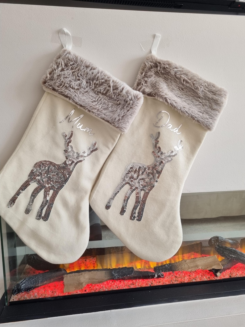 May include: Two cream-colored Christmas stockings with faux fur trim and a silver sequin reindeer design. One stocking is personalized with "Mum" and the other with "Dad".