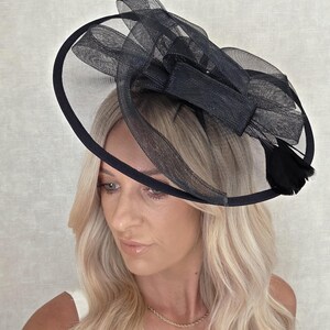 May include: A black formal hat with a circular brim and a mesh overlay. The hat features a large bow and feather accents. The hat is designed for special occasions.