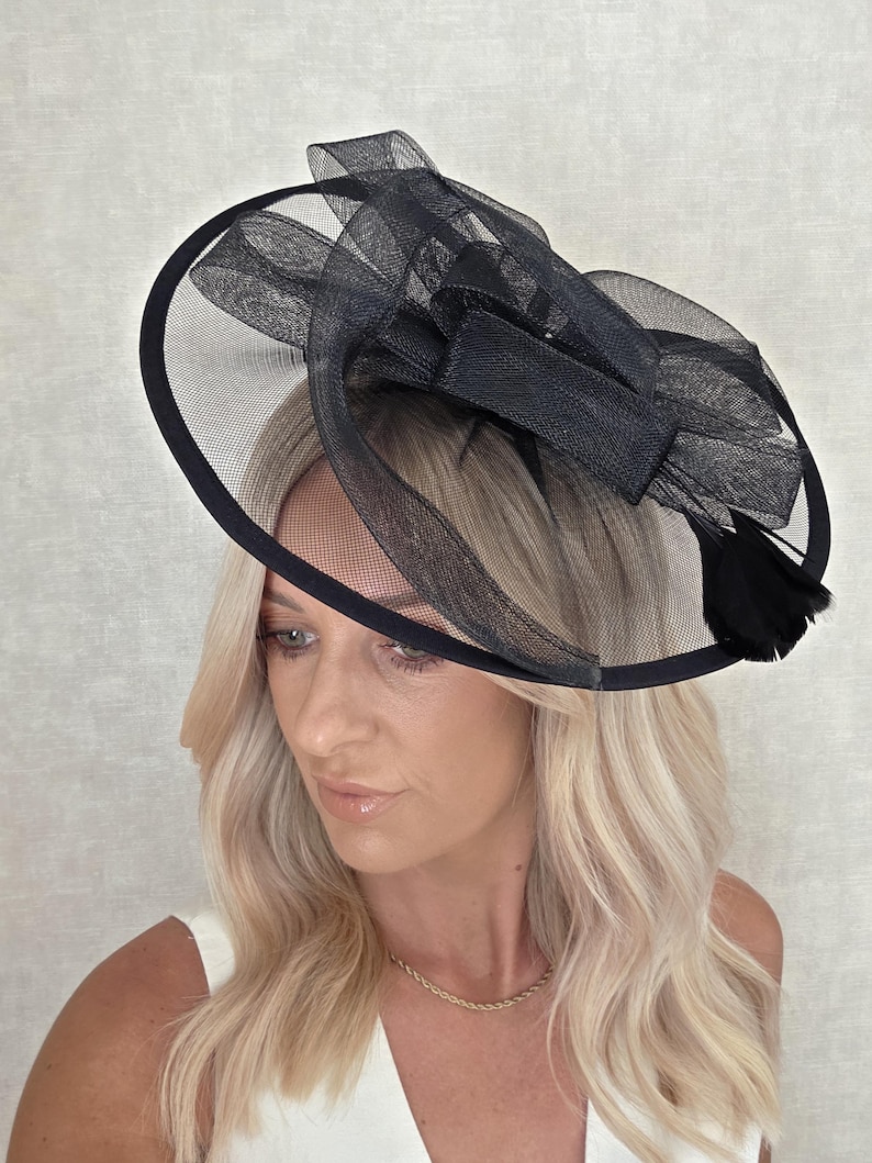 May include: A black formal hat with a circular brim and a mesh overlay. The hat features a large bow and feather accents. The hat is designed for special occasions.