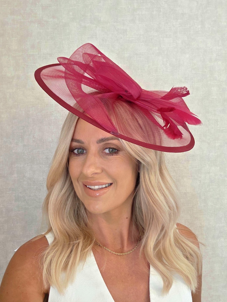 May include: A magenta-colored hat with a wide brim and a decorative bow made of sheer fabric. The hat features a dark red trim and is adorned with feathers. This formal headwear is suitable for special occasions.