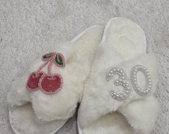 May include: A pair of white, fluffy slippers. One slipper features a beaded cherry design, while the other has the number "30" embellished with pearls. The slippers have a soft, plush texture.
