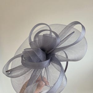 May include: A gray fascinator with a circular mesh base and decorative loops. The headpiece features a central cluster of fabric and a thin headband. Feathers are attached to the base. This formal headwear is suitable for special occasions.