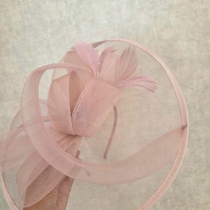 May include: A pale pink fascinator with a circular mesh base and a decorative bow. The bow is made of sheer fabric and adorned with delicate feathers. The headpiece is designed for special occasions.