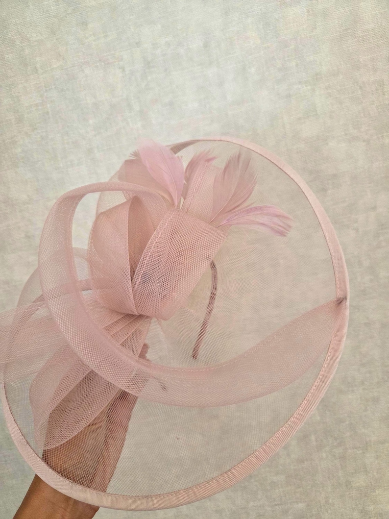 May include: A pale pink fascinator with a circular mesh base and a decorative bow. The bow is made of sheer fabric and adorned with delicate feathers. The headpiece is designed for special occasions.