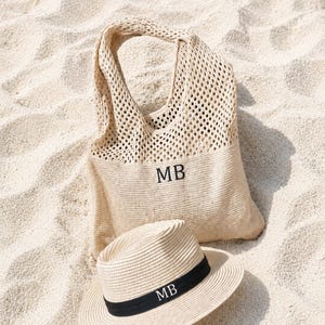 May include: A beige tote bag and a straw hat with a black band, both with the initials "MB" embroidered on them, rest on a sandy surface. The bag has a woven design, and the hat has a classic fedora shape.