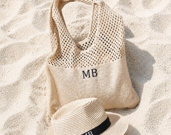 May include: A beige tote bag and a straw hat with a black band, both with the initials "MB" embroidered on them, rest on a sandy surface. The bag has a woven design, and the hat has a classic fedora shape.