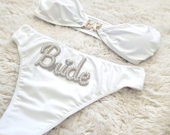 May include: White bridal bikini with a pearl embellished "Bride" on the bottom piece and a white bandeau top with a rhinestone embellishment.