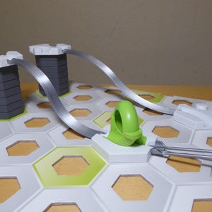Puede incluir: Un colorido juego de juguetes de circuito de canicas. El juego incluye torres grises con tapas blancas, pistas plateadas y un bucle verde. La base es blanca con patrones hexagonales verdes y amarillos.