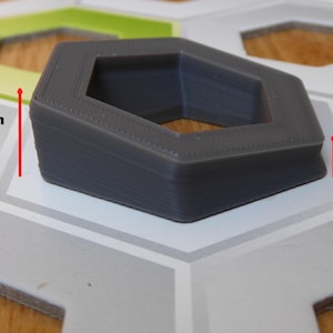 May include: A gray, hexagonal-shaped object, 1.5 cm tall and 0.5 cm thick, sits on a patterned surface. The object has a hollow center and appears to be 3D-printed. The design is geometric and modern, suitable for various applications.