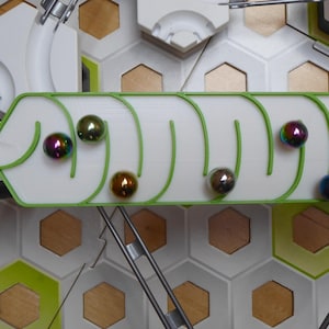 May include: A marble run game with a white track outlined in green, featuring several colourful marbles. The track is set against a backdrop of white and gold hexagonal tiles, with metal support structures.