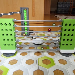 May include: A marble run construction set with green supports and silver rails. The set includes a spiral lift and various colored marbles. The base is a hexagonal tile pattern. The set is designed for building and playing.