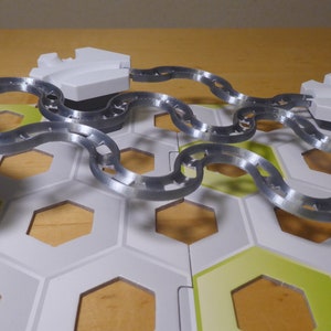 Puede incluir: Primer plano de un juego de mesa con una cadena plateada que conecta dos piezas hexagonales blancas. El tablero est&aacute; hecho de baldosas hexagonales blancas y verdes.