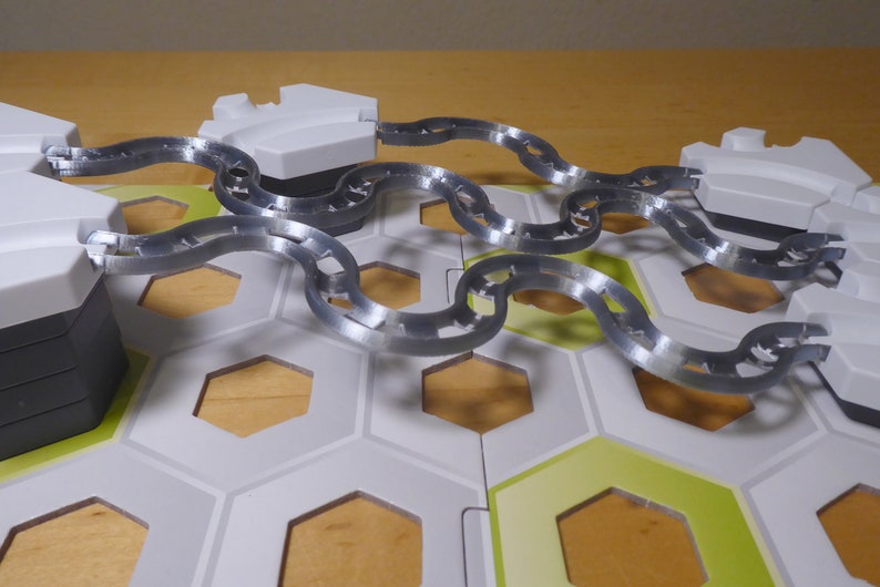 Puede incluir: Primer plano de un juego de mesa con una cadena plateada que conecta dos piezas hexagonales blancas. El tablero est&aacute; hecho de baldosas hexagonales blancas y verdes.