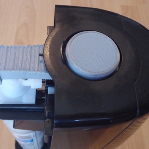 May include: A black and gray water dispenser with a white plastic top and a gray filter. The filter is visible through a round opening in the top of the dispenser.