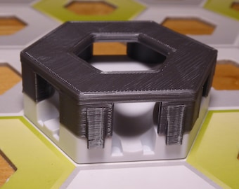Puede incluir: Una pieza de juego hexagonal gris oscuro con un centro hueco y cuatro patas. La pieza está sentada en una baldosa hexagonal blanca.
