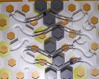 Pode incluir: Um jogo de mármore com azulejos hexagonais cinzentos e brancos e trilhos prateados. Existem quatro mármore no jogo, um preto, um roxo, um dourado e um azul.