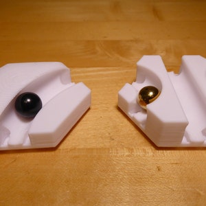 Op de afbeelding: Twee witte, 3D-geprinte plastic objecten met een groef in het midden. Een object heeft een zwarte bal in de groef en het andere heeft een gouden bal in de groef.