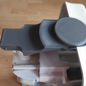 May include: A grey plastic 3D printed part with a round knob on the end. The part is attached to a white plastic base.