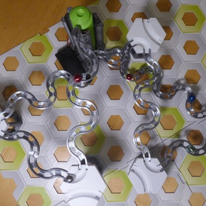 Puede incluir: Una pista de canicas de plata con azulejos hexagonales blancos y detalles verdes. La pista est&aacute; montada sobre una superficie de madera e incluye tres canicas rojas, una canica azul y una canica plateada.