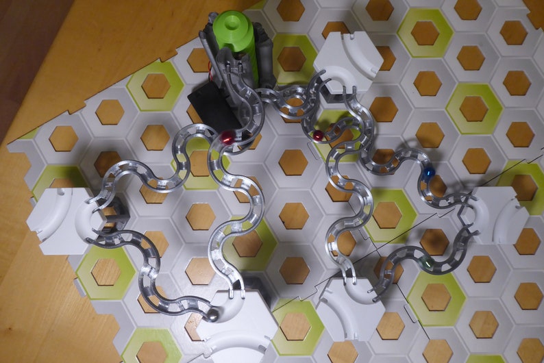 Puede incluir: Una pista de canicas de plata con azulejos hexagonales blancos y detalles verdes. La pista est&aacute; montada sobre una superficie de madera e incluye tres canicas rojas, una canica azul y una canica plateada.