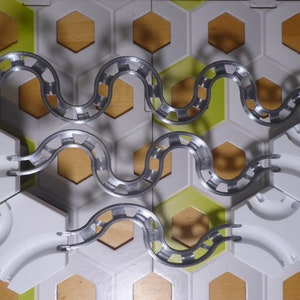 Puede incluir: Primer plano de un juego de canicas de plata con baldosas hexagonales blancas y fondo de madera. El juego est&aacute; configurado con una pista sinuosa que sube y baja.