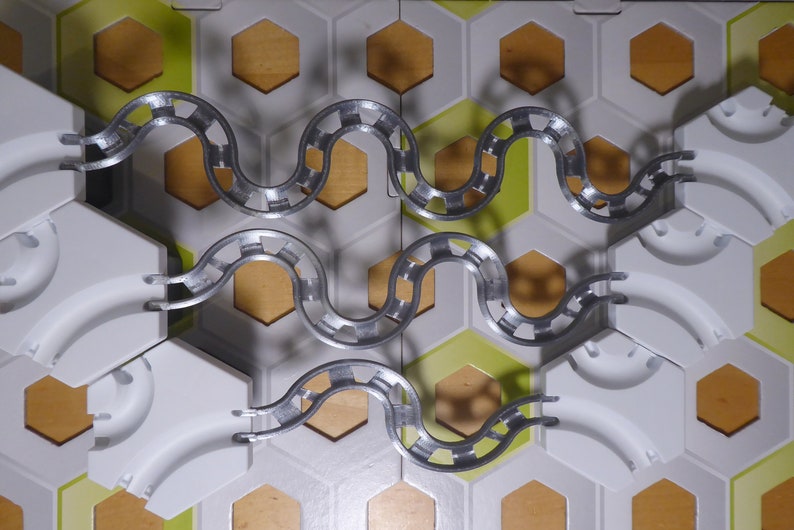 Puede incluir: Primer plano de un juego de canicas de plata con baldosas hexagonales blancas y fondo de madera. El juego est&aacute; configurado con una pista sinuosa que sube y baja.